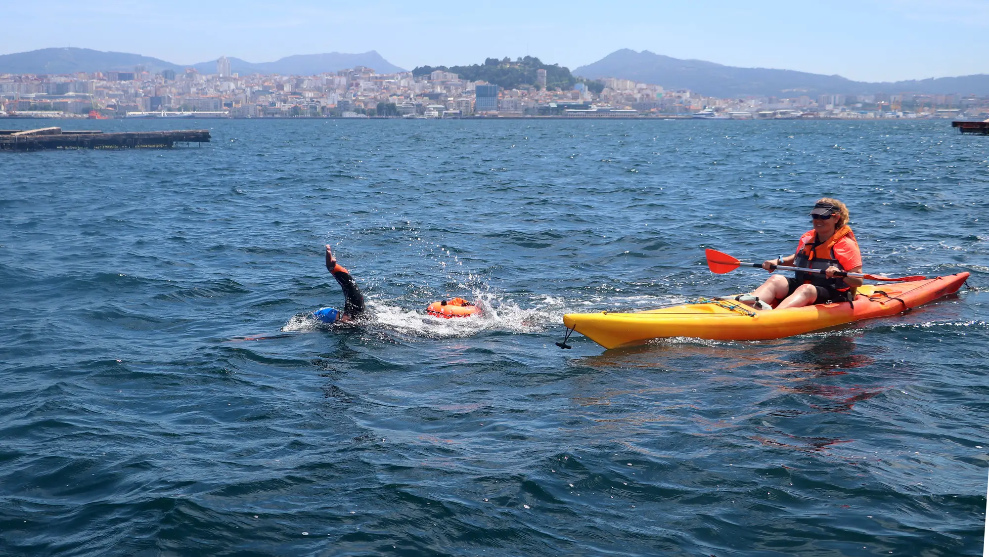 Un nadador avanza a crol junto a un kayak de apoyo, con la ciudad de Vigo al fondo